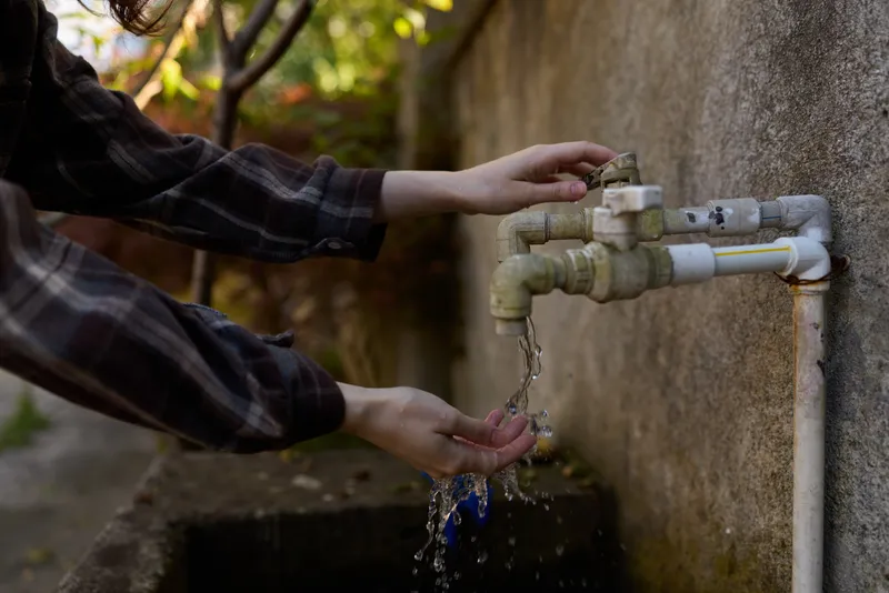 A young woman wearing a plaid shirt washes her hands under a weathered outdoor faucet, water streaming from the spout while soft sunlight filters through leaves, creating a calm everyday moment