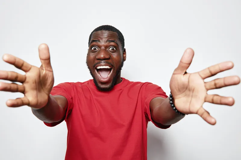 African American man with dark skin wearing red t shirt smiling widely and reaching out with open hands isolated on white background for happiness and excitement concept.