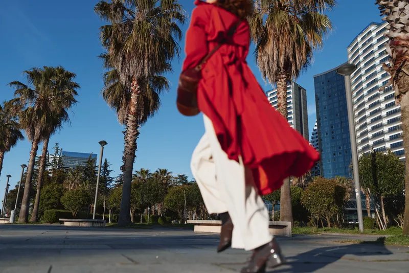 Woman in a red coat walks along a sunny urban street, outdoor fashion scene with palm trees and modern buildings, sleek casual wear, dynamic movement and confident mood