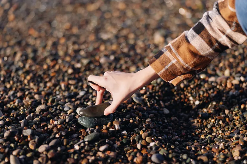Closeup of a hand picking a dark stone from a bed of pebbles on a beach, showcasing natural texture and tactile detail in an outdoor setting.