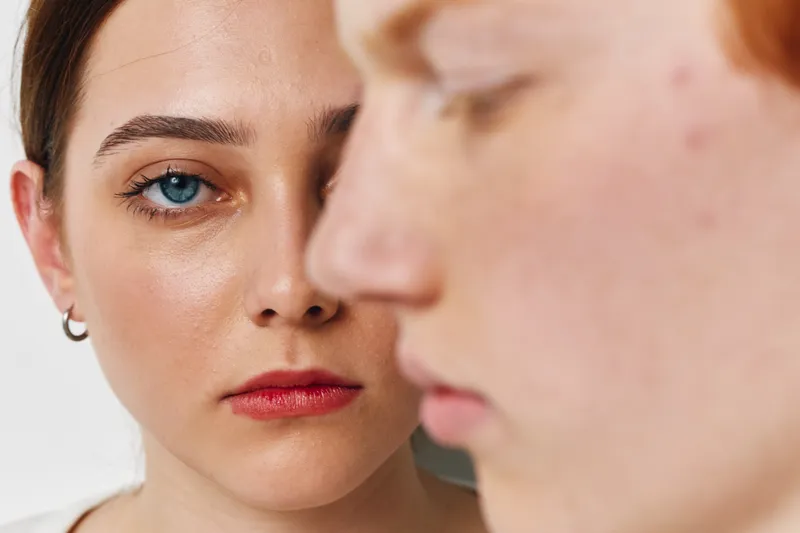 Portrait of woman with blue eye and red lipstick looking directly at camera behind blurred profile of another woman. Closeup of female faces with natural skin and emotional expression.