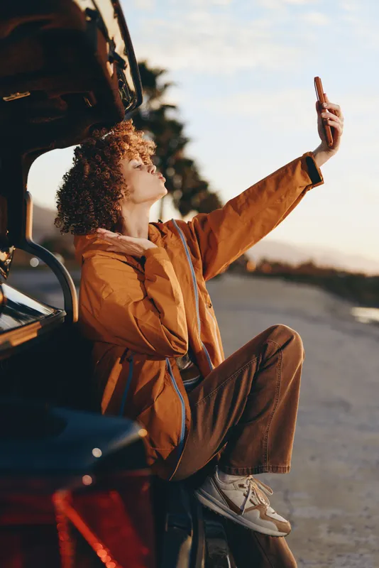 Woman lifestyle on road at sunset, sitting in car door open, waving hand, curly hair, brown fashion jacket and jeans, travel outdoors, warm golden light
