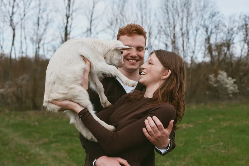 A joyful couple laughs as they cradle a playful goat kid outdoors, sharing affection in a rural setting. A candid lifestyle moment showing companionship, warmth, and animal interaction in a bright,