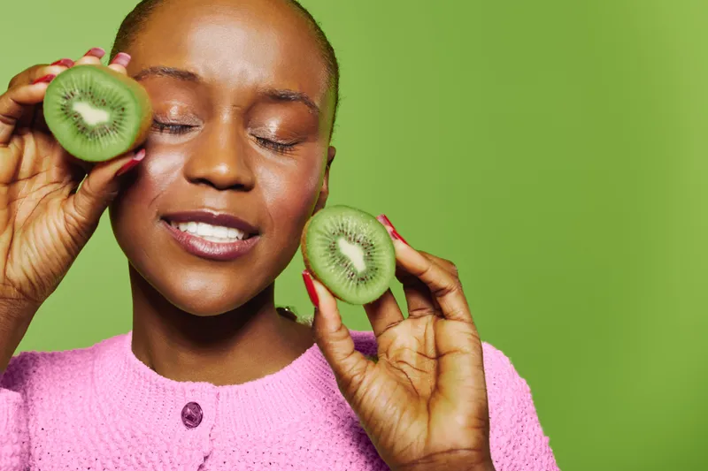 Happy African woman with dark skin holds two kiwi halves near her face, eyes closed and smiling. The vibrant green background highlights the fresh fruit and joyful mood of wellness and health.
