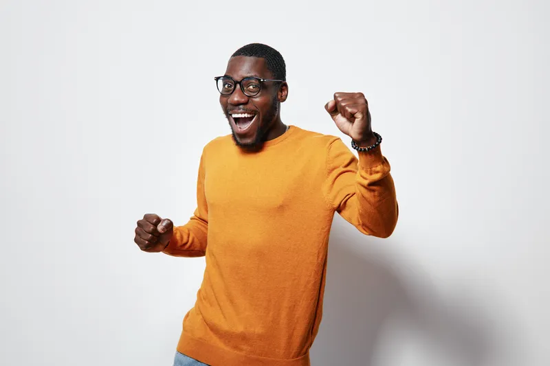 African American man with dark skin wearing orange sweater and glasses smiling and celebrating with fists up on white background in studio.