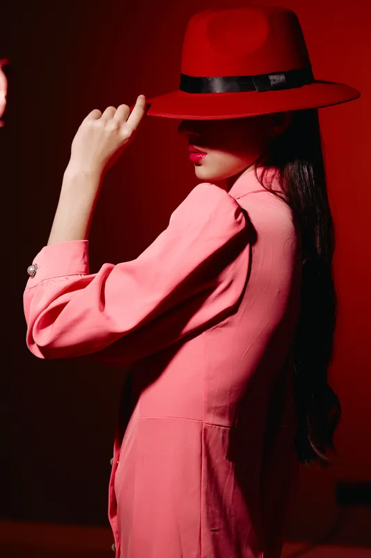 Woman with long dark hair wearing a pink jacket and a wide-brimmed red hat covers her face with one hand. Studio shot with vibrant red background highlights fashion and beauty creativity.