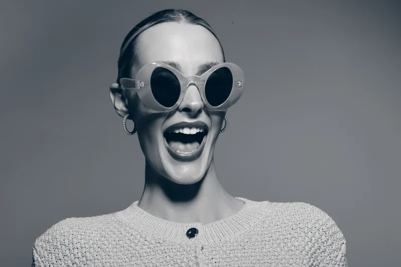 Happy young white woman with pale skin wearing round sunglasses and earrings smiles widely. Studio portrait with bright lighting and casual knitwear, fashion and lifestyle concept.