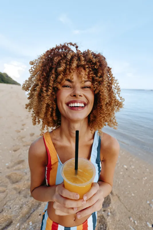 Woman with curly hair smiling on a beach, holding a coconut drink with a straw. Ocean and sky behind, warm sunlight, summer vacation mood, relaxed outdoors today.