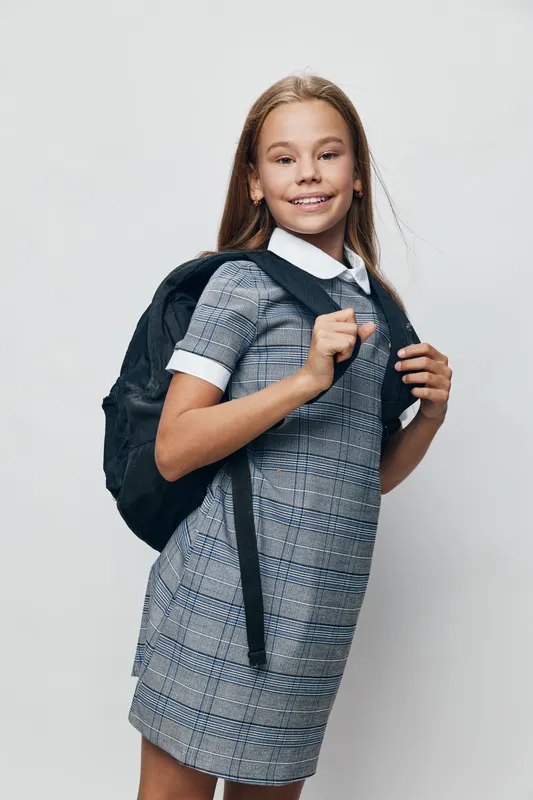 Schoolgirl with long hair wearing checkered dress and white collar smiling, holding black backpack straps on neutral background, posing for school portrait or education concept.