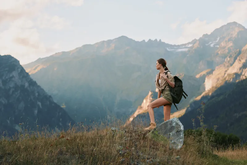 Woman hiker with backpack stands on a rocky ledge in the mountains, embracing outdoor travel and nature, capturing a moment of adventure against vast alpine panorama