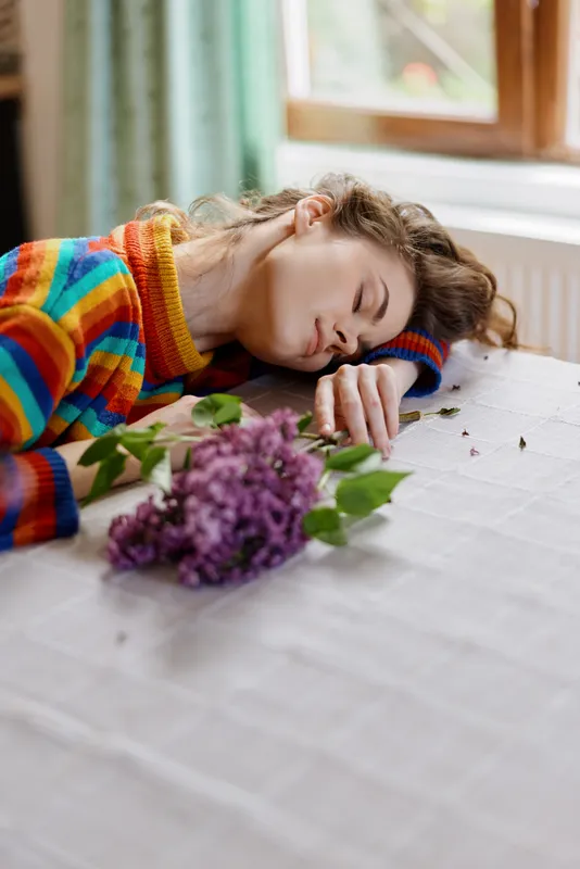 Young woman rests her head on a table beside a lilac bouquet, wearing a colorful striped sweater, in a sunlit living space, expressing calm domestic mood and rest.