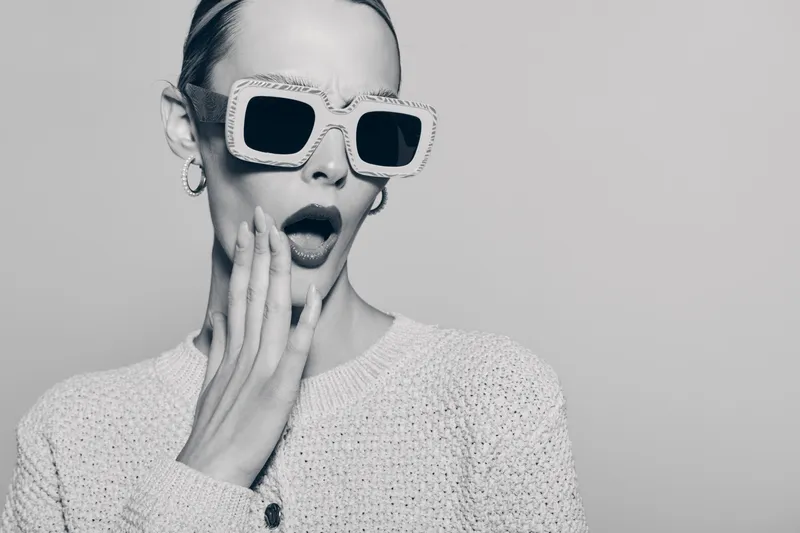 Surprised white woman with pale skin wears large stylish sunglasses and knitted sweater, touching cheek with open mouth. Concept of fashion and expression.