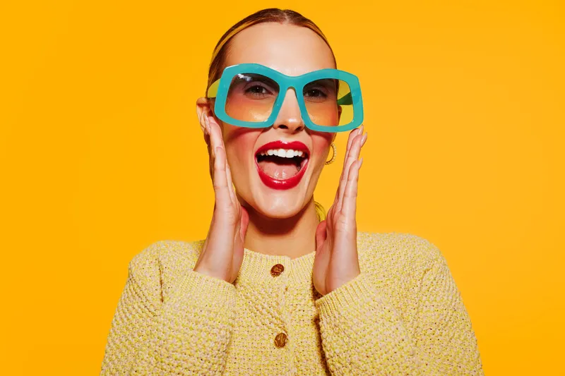 Excited woman in large blue sunglasses and yellow sweater smiles widely on bright orange background. Concept of happiness, fashion, summer style, and joyful emotions.