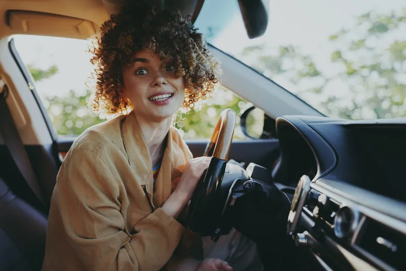 Smiling woman with curly hair sits in a car, close-up on the steering wheel, warm retro film colors illuminate a lifestyle moment of travel and joy.
