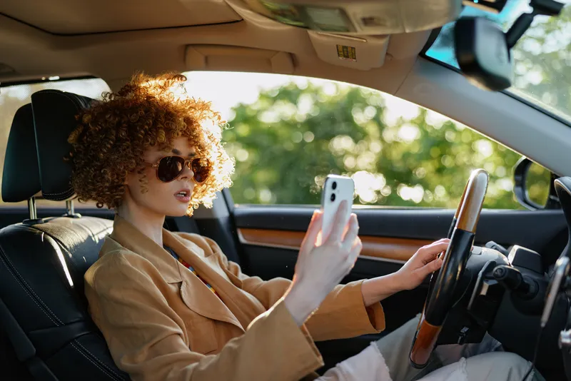 Curly haired woman wearing sunglasses sits in a car, looking at a smartphone. Natural daylight highlights casual fashion and lifestyle, with a warm modern interior vibe.