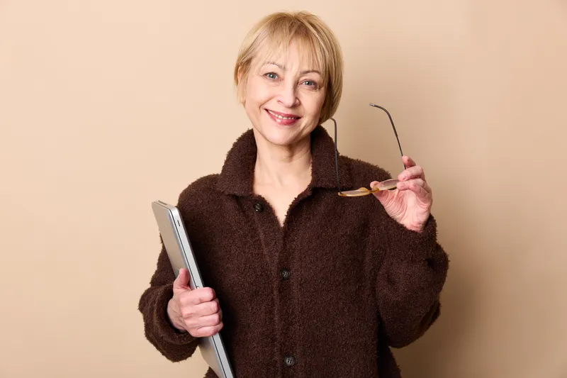 Woman elderly glasses smiling portrait beige studio holding fur coat, friendly senior holds reading eyewear, relaxed pose, calm indoor background copyspace