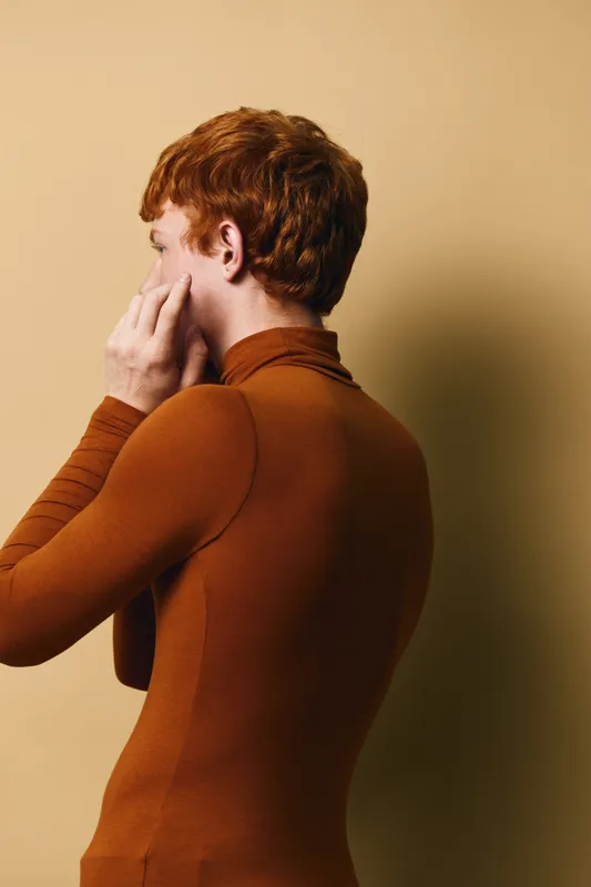 Redhead man in brown turtleneck sweater posing sideways with hand on face against beige background. Portrait of young male model in casual autumn clothing with thoughtful expression.