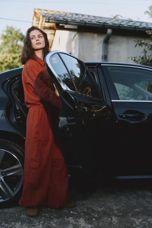 A woman in a red jumpsuit steps from a sleek car, posing in a driveway, embodying everyday confidence and contemporary fashion in a casual lifestyle moment