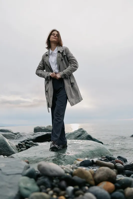 Young woman in a patterned coat stands confidently on rocky shore near sea under cloudy sky. Casual fashion and nature concept with calm mood and outdoor lifestyle by the water.