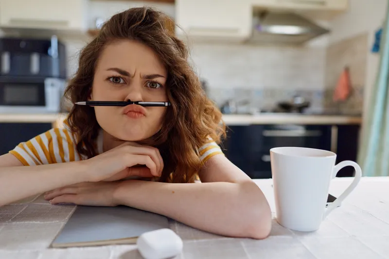 Woman lifestyle at home sits at a kitchen table with a pen in her mouth, frustrated expression, working from home, papers and mug scattered on the surface, candid domestic scene