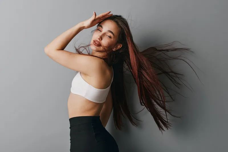 Young woman with long flowing hair posing in white top and black pants against gray background. Fashion portrait with motion, beauty, elegance, and casual style in studio lighting.