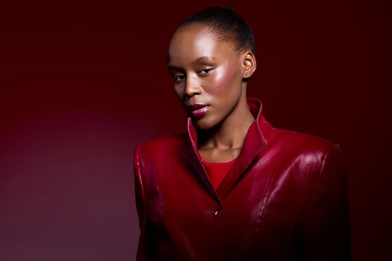 Portrait of confident African woman with dark skin wearing shiny red jacket on red background. Studio lighting highlights her smooth skin and elegant expression. Fashion concept.