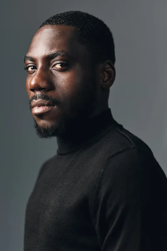 African American man with dark skin wearing black turtleneck sweater looking at camera in creative visual designer style portrait with studio lighting and neutral background.