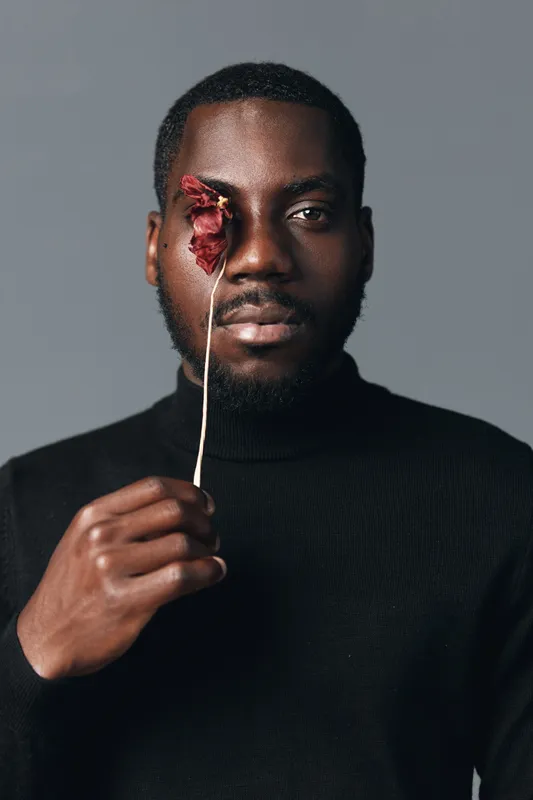 African man with dark skin holding dried red flower in front of eye in creative conceptual visual style. Portrait with black turtleneck and neutral background for artistic expression.
