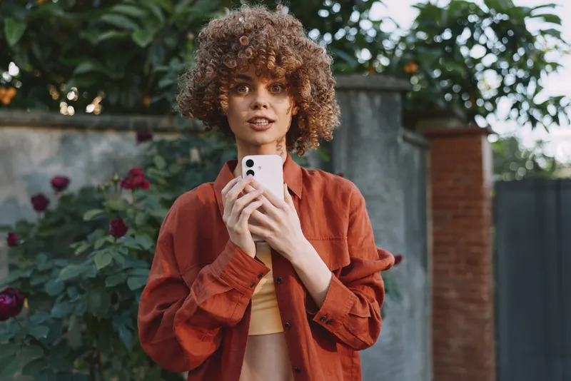 Young woman with curly hair holds a smartphone outdoors, wearing a rust jacket and beige top, looking at the camera with a relaxed, confident vibe