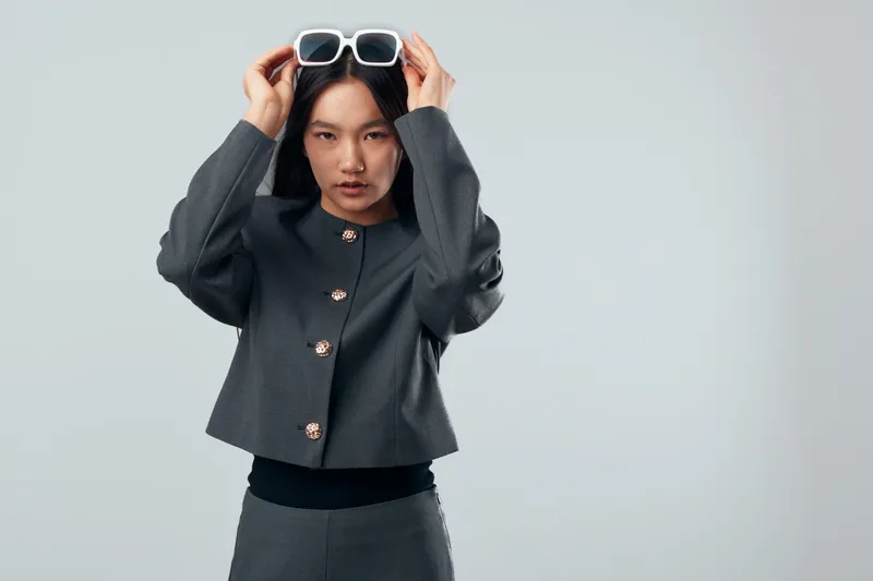 Woman with straight black hair wearing gray jacket and pants adjusts white sunglasses on head against plain background. Fashionable young model posing with focused expression indoors.