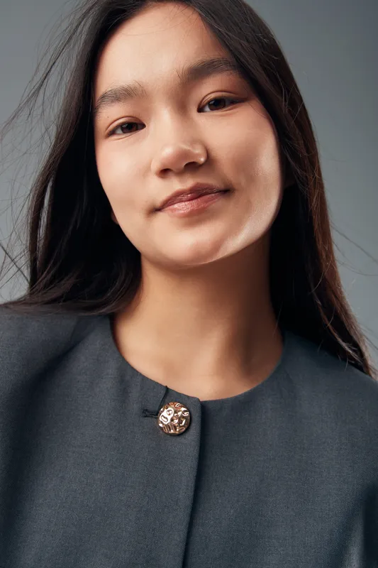 Portrait of young woman with long black hair wearing dark jacket with decorative button looking at camera. Closeup face expression with smooth skin and natural makeup in studio light.