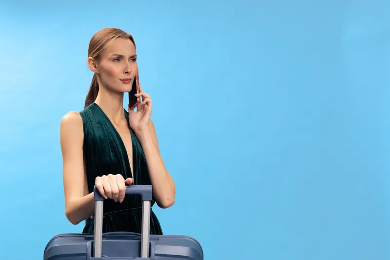 Young woman with suitcase talking on smartphone against blue background. Business travel concept with female traveler dressed in elegant outfit ready for trip.