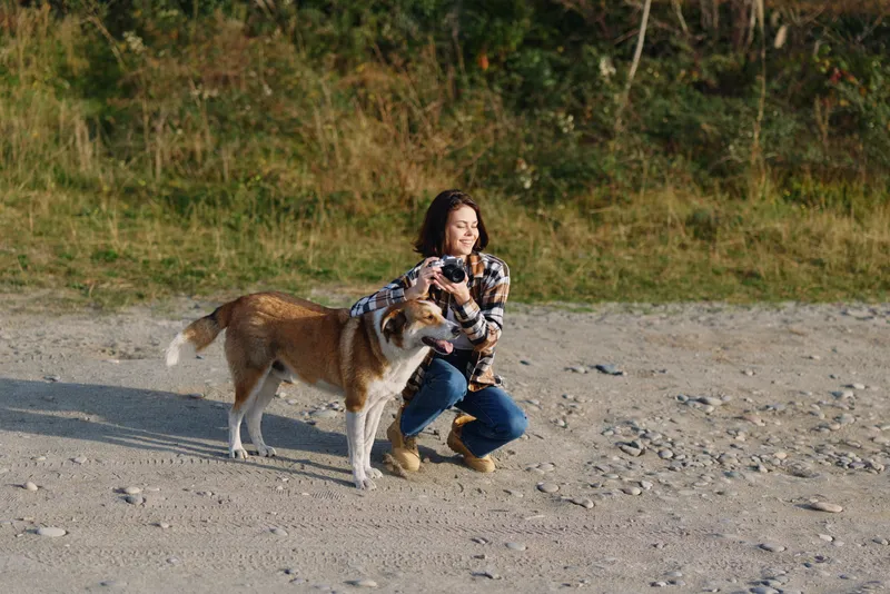 Woman crouching on a sandy beach embraces two dogs, enjoying a joyful outdoor moment with a grassy backdrop and clear sky, capturing affection, energy and a peaceful bonding scene