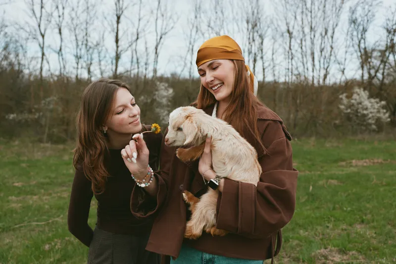 Cheerful outdoor lifestyle scene featuring two women with a playful goat kid, sharing smiles and gentle interaction in a sunlit grassy field, rustic outfits, natural light, warm mood, authentic