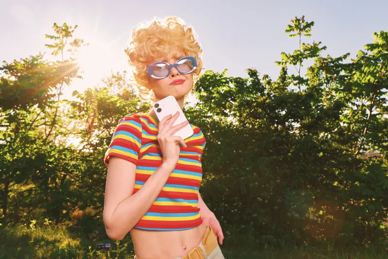 Smiling woman with curly hair in a striped top outdoors during golden hour. Sunny park scene with retro vibe, sunglasses, and a handheld phone. Crisp light enhances playful mood and casual lifestyle