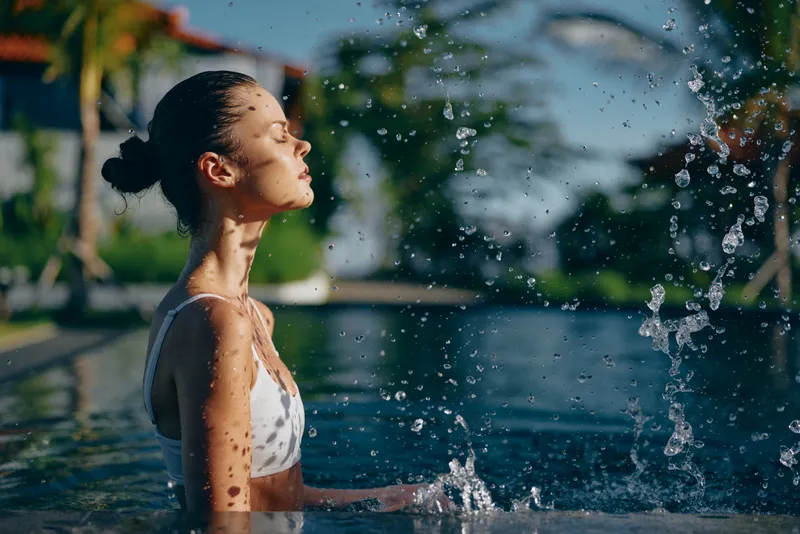 A woman stands in a bright pool as water sprays around, soaking up sun, shoulders sunlit, hair in bun, white swimsuit. Tropical backdrop, droplets glisten, conveying relaxation, outdoor leisure,