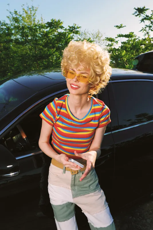 Smiling woman with curly blonde hair wearing a striped rainbow top and colorblocked jeans beside a dark car. Retro street style, sunny vibe, playful confidence, bold colors.