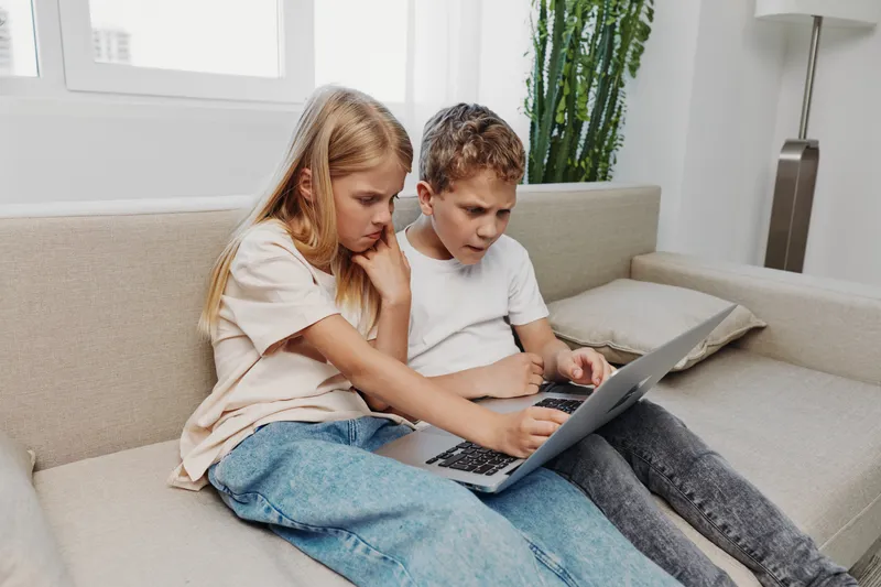 Two focused children sitting on a sofa using a laptop together in a bright modern room, concentrating on the screen. Concept of learning, technology, and teamwork.