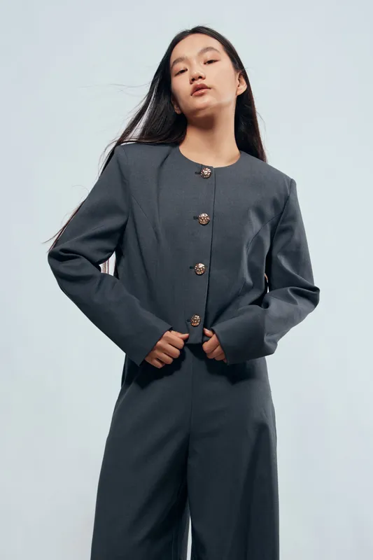 Young woman posing in grey tailored suit with long sleeves and button details standing confidently against light background in studio. Fashion, style, elegance and modern clothing.
