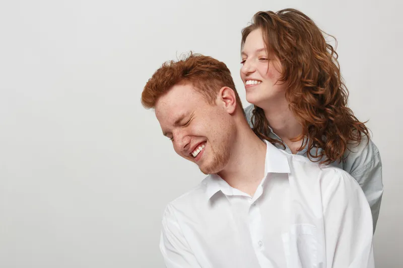 Happy young couple with red hair and curly hair laughing together on light background. Concept of love, friendship, joy, and positive emotions in casual clothing and natural style.