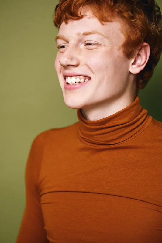 Young man with red hair and freckles wearing brown turtleneck smiling and looking sideways on green background. Portrait of cheerful male with natural skin and casual outfit.