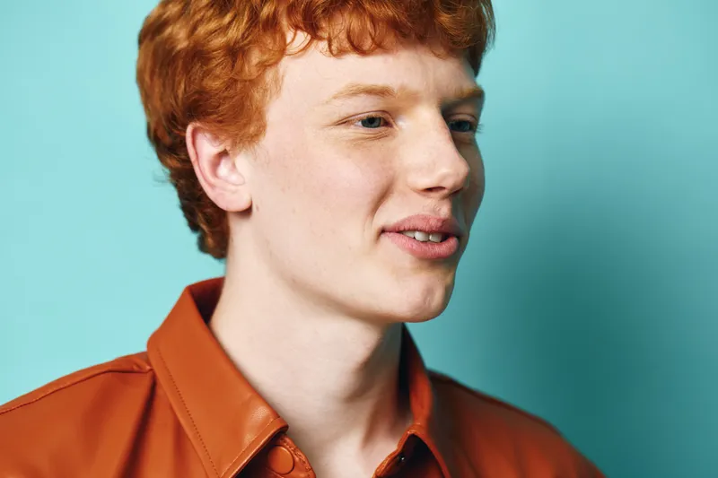 Young man with red hair and freckles wearing brown leather jacket smiling and looking sideways against blue background. Portrait, casual style and natural expression in studio lighting.