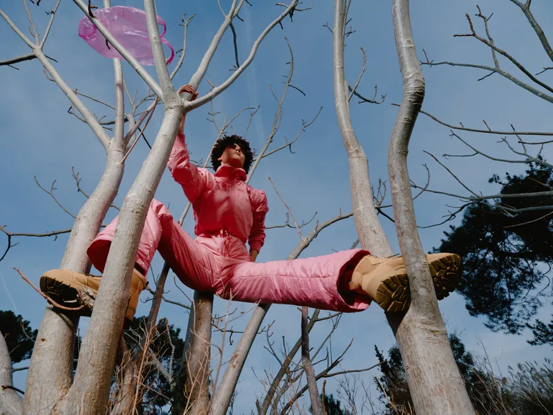 Acrobat, tree, outdoor, performance, balance, costume, pink jumpsuit high among bare branches against blue sky, daring pose, athletic display, vibrant color, playful stunt, nature scene