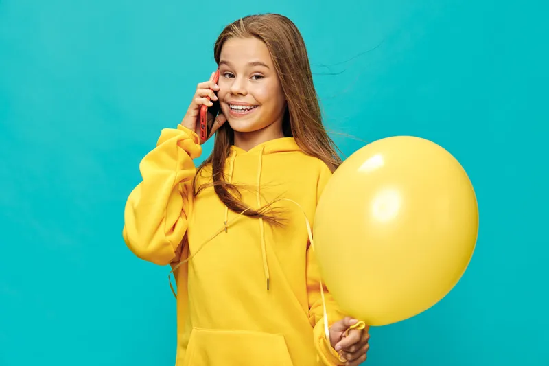 Teenage girl in yellow hoodie talking on smartphone and holding yellow balloon with smile on blue background, casual style, young female communication and happiness concept.