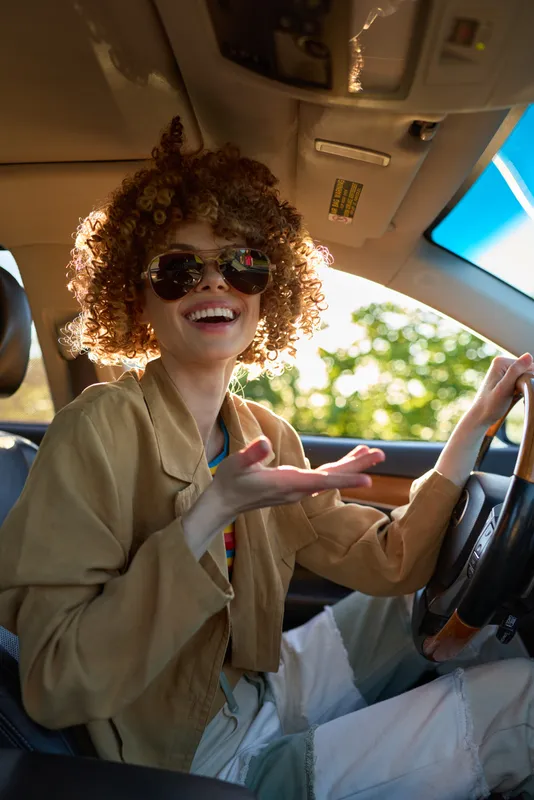 Vibrant woman driving a car, cheerful smile, sunglasses reflecting sunlight, casual blazer, warm beige interior, outdoors visible through window, energetic lifestyle moment in bright light