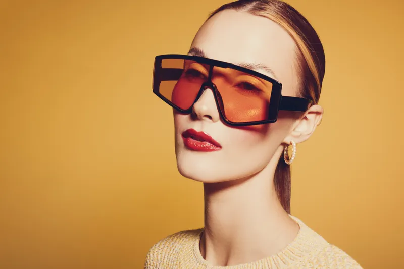Stylish woman with oversized orange sunglasses and red lipstick poses against yellow background. Fashion portrait with modern eyewear and confident expression.
