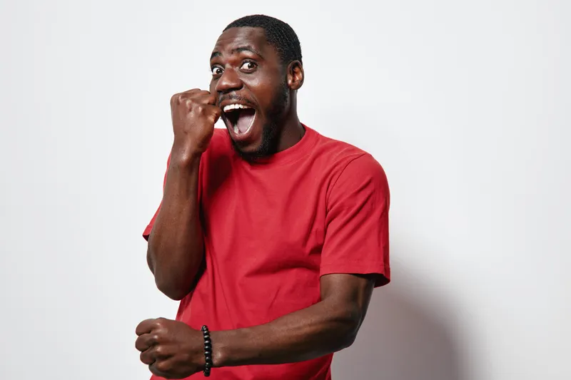 African American man with dark skin wearing red t shirt showing excitement and joy with clenched fist and open mouth on white background. Happy male expressing success and celebration.