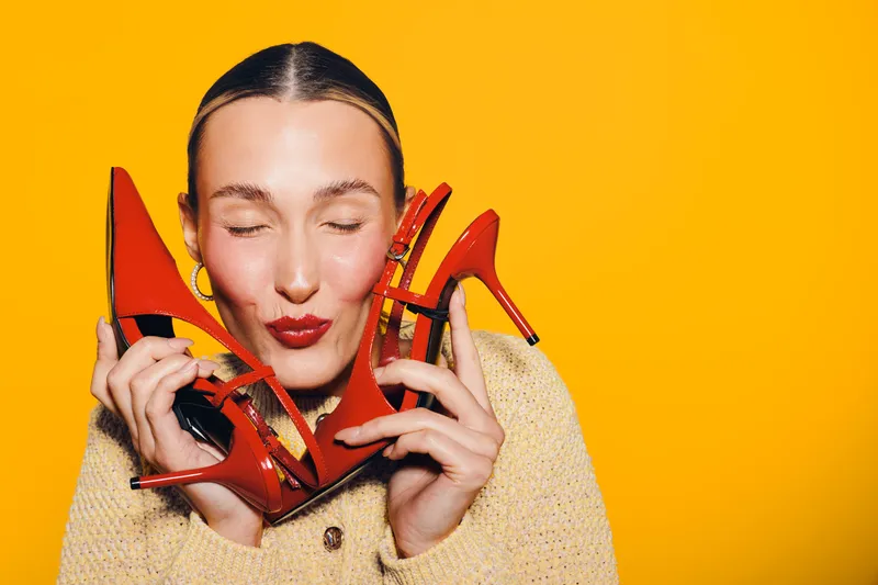Young woman with closed eyes holds red high heel shoes near face, smiling with red lipstick on bright yellow background. Concept of fashion, style, and joyful emotions.