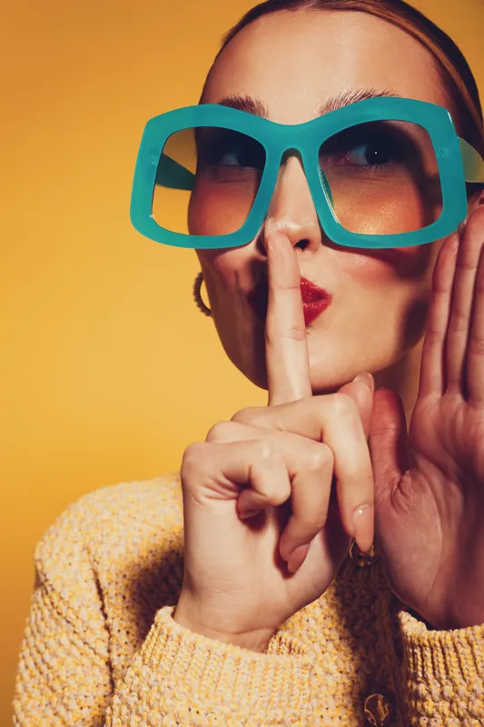 Woman in large blue glasses and yellow sweater makes shushing gesture with finger on lips against orange background. Concept for secrecy, silence, and quiet communication.