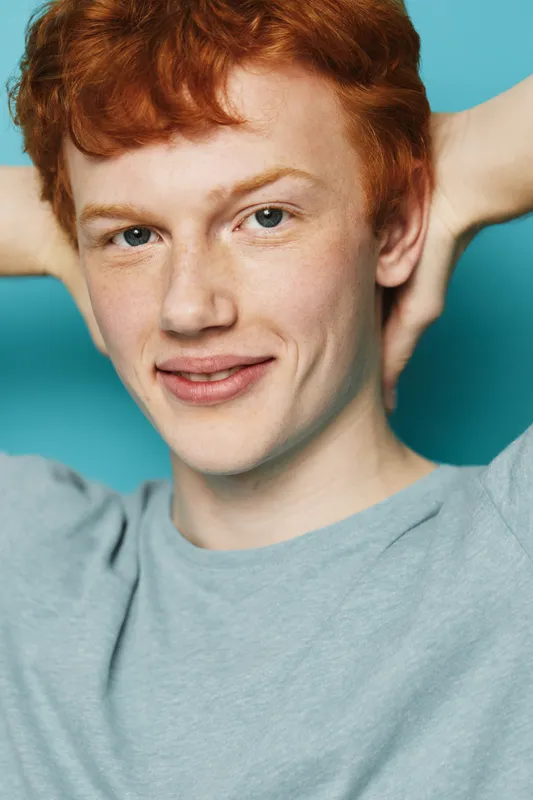 Young man with red hair and freckles smiling with hands behind head, wearing grey shirt against blue background. Casual portrait of confident male with relaxed posture and friendly expression.